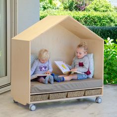 two young children sitting in a wooden house bed reading books on the floor outside with plants and bushes behind them