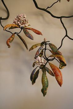 a metal branch with flowers and leaves hanging from it's center, in front of a white wall