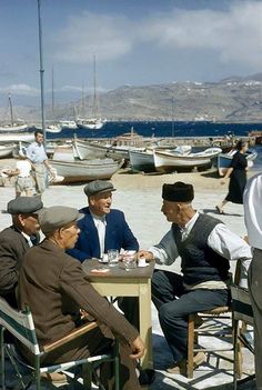 four men sitting at a table on the beach with boats in the water behind them