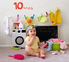 a baby sitting on the floor in front of a chalkboard with clothes hanging from it