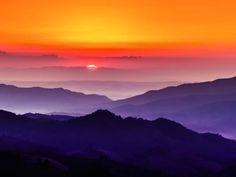 the sun is setting over some mountains in the foggy morning sky, with purple and orange hues