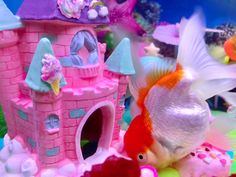 there is a goldfish in front of a pink castle and other toys on the seabed