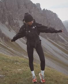 a woman standing on top of a grass covered hillside next to a mountain range with her arms outstretched