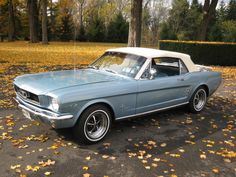 an old blue mustang parked in a parking lot with autumn leaves on the ground and trees