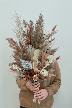 a woman holding a bouquet of dried flowers in her hands and wearing a brown sweater