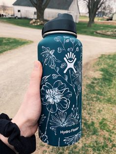 a person holding up a hydro flask in front of a dirt road and house