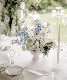 a table set with white and blue flowers in a vase, silverware and candles