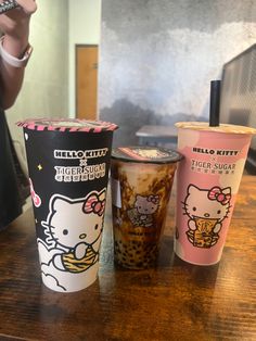 three hello kitty cups sitting on top of a wooden table next to a person holding a cell phone