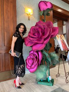 a woman standing next to a large pink rose sculpture in front of a wall with paintings on it