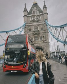 a red double decker bus driving past a tall building with a bridge in the background