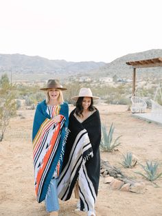 two women in hats and scarves are standing on the desert with their arms around each other