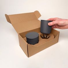 a hand is holding a cup in a cardboard box with two cups inside the box