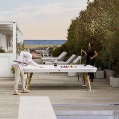 a man and woman playing pool on the deck of an oceanfront hotel with lounge chairs in the background