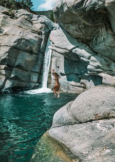 a person jumping into a body of water next to some large rocks and boulders with a waterfall in the background