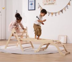 two young children playing on a wooden bed frame in a room with white walls and wood flooring