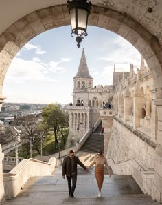 two people holding hands while walking up some stairs in front of a castle like building