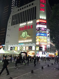 many people are walking around in front of a large building with neon signs on it