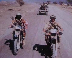 two men riding motorcycles down a dirt road in the middle of the desert with no one on them