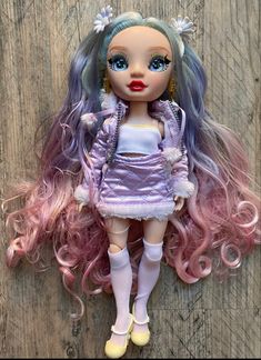 a doll with pink and purple hair is laying on a wooden floor next to a wall