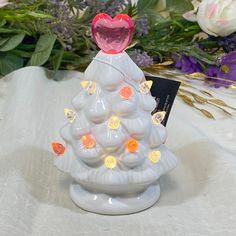 a small white ceramic christmas tree with lights on it's base and flowers in the background