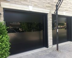 two black garage doors are open in front of a brick building with green bushes on the sidewalk