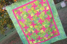 a green and pink quilt hanging from a clothes line in the grass with trees in the background