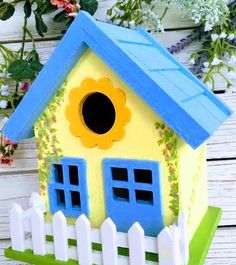 a yellow birdhouse with blue windows and a white picket fence in front of flowers