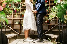 a man and woman standing next to each other in front of a plant wall with flowers on it