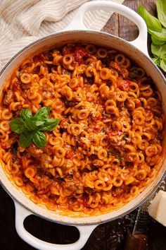 a large pot filled with pasta and sauce on top of a wooden table next to basil leaves