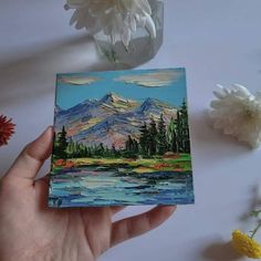 a hand holding up a small card with mountains in the background and water below it