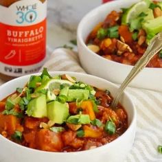 two white bowls filled with chili and avocado next to a bottle of beer