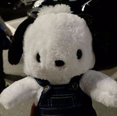 a white teddy bear with black eyes and overalls on it's chest, sitting in front of a car