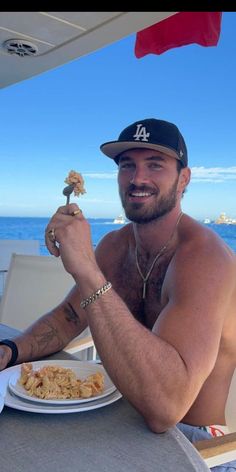 a shirtless man sitting at a table with food in front of him and the ocean behind him