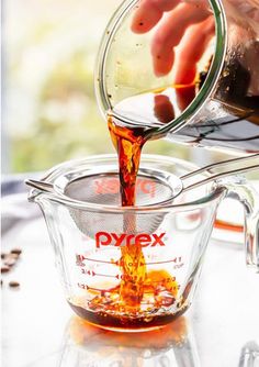 a person pouring syrup into a measuring cup on top of a glass table next to coffee beans