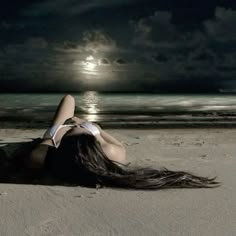 a woman laying on top of a sandy beach next to the ocean under a full moon