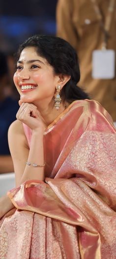 a woman in a pink sari smiles at the camera while sitting down with her hand on her chin