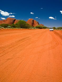 a white car driving down a dirt road in the desert with red rocks behind it