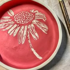 a red plate with a flower painted on it next to some paintbrushes and a brush