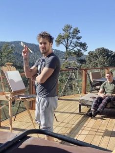 a man standing on top of a wooden deck holding a paintbrush and an easel