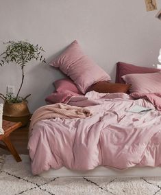 an unmade bed with pink linens and pillows in a white room next to a potted plant