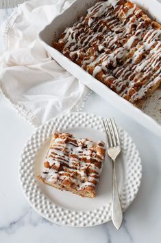 a white plate topped with a piece of cake next to a pan filled with cinnamon rolls