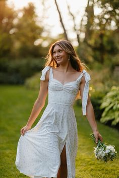 a woman in a white dress holding a bouquet and walking through the grass with her legs crossed