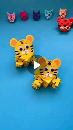 two paper toy tigers sitting next to each other on a blue surface with cats in the background