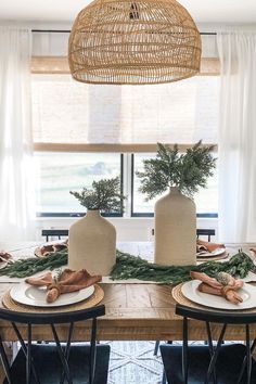 the table is set with two vases and greenery on top of it, along with other place settings