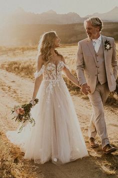 a bride and groom holding hands while walking down a dirt road in the desert at sunset