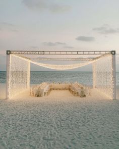 an outdoor wedding set up on the beach with white linens and lights strung over it