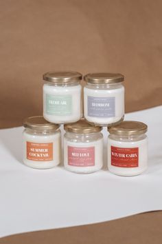 five jars of candles sitting on top of a white tablecloth covered table with a brown background