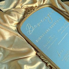 a blue and gold wedding card laying on top of a satin covered bed sheet with the word bevergas written in cursive writing