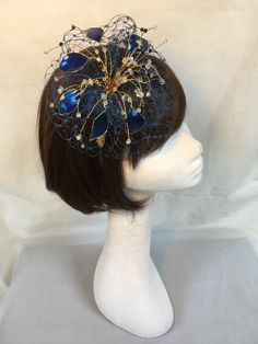 Delicate gold and blue fascinator perfect for the guest who wants something fairly understated. This design is a beautiful selection of glass effect leaves and flowers clustered together amongst a fine layer of veiling and set onto a fine comfort base tiara band. As with all our designs, this piece can be made exact to the image or in any colour combination to match in with your outfit. If a specific colour is required customers can message over an image of their outfit and accessories, or if ti Blue Structured Mini Hat For Wedding, Blue Adjustable Fascinator For Gift, Adjustable Blue Headpieces For Party, Blue Headband For Party, Elegant Blue Headband As Gift, Blue Headband Party Costume Hat, Elegant Blue Headpiece For Events, Blue Party Headband, Elegant Blue Hair Accessories For Party