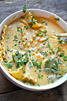 a white bowl filled with pasta and greens on top of a wooden table next to a spoon